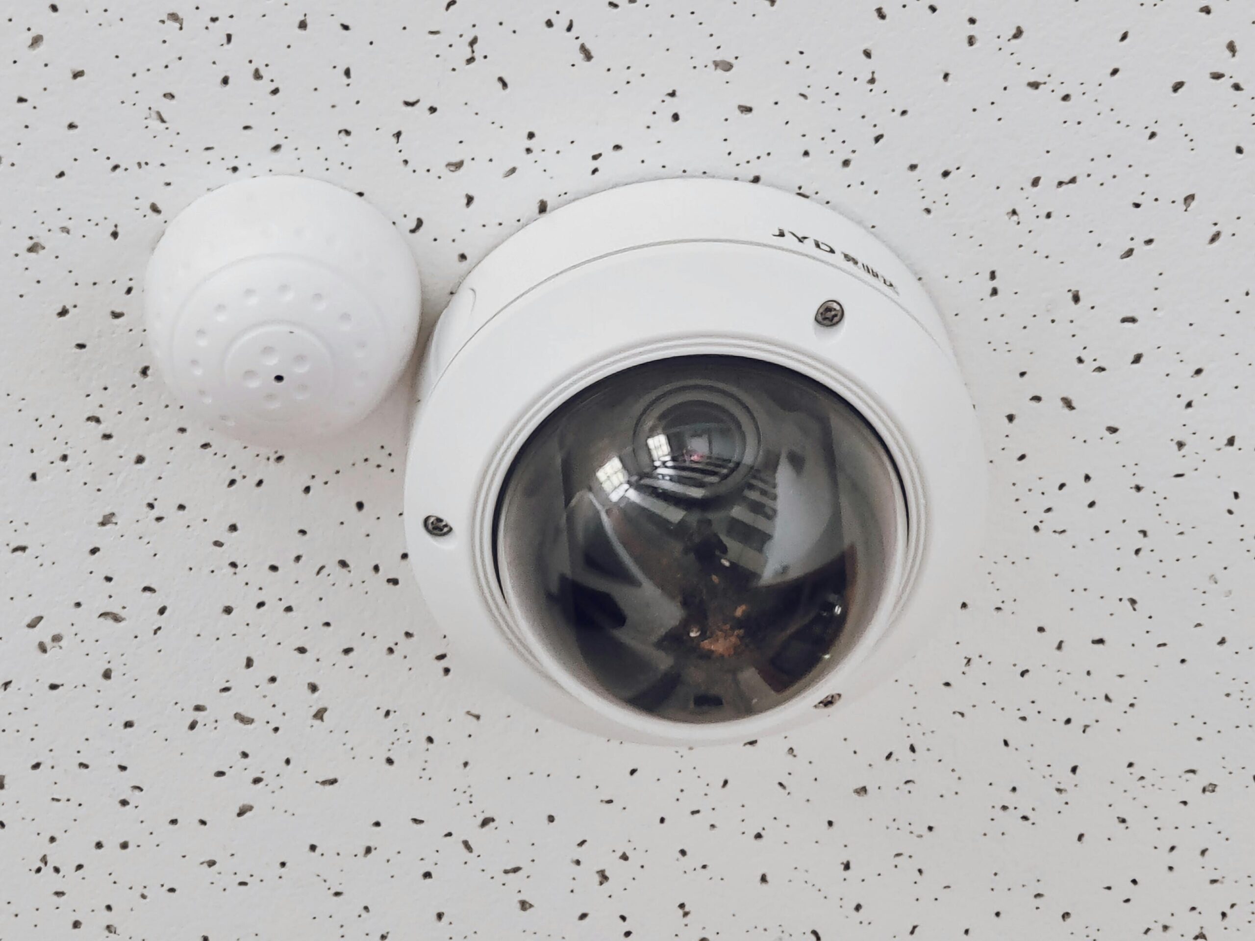 Home Page Close-up of a round dome surveillance camera installed on a speckled ceiling for security monitoring.